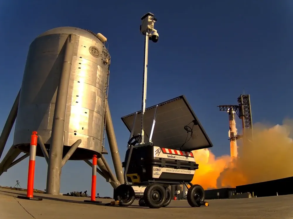 A LOBO tower at a launch site in South Texas, mast up, with a rocket lifting off behind it.