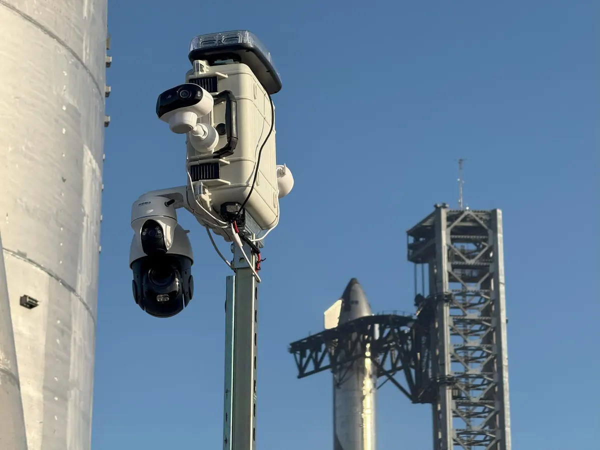 The camera head at the top of LOBO's mast, with a rocket on the launch tower behind it.