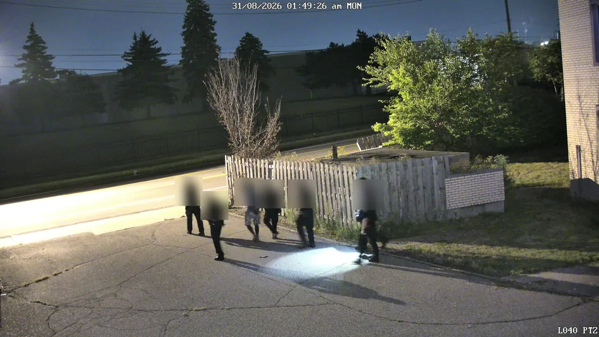 Tower camera at night: police officers walk a person out along a wooden fence beside the building.