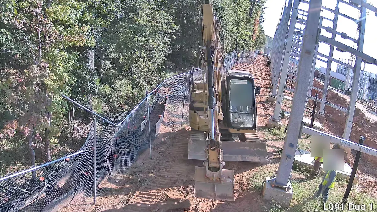 Tower camera: an excavator parked against a newly built chain-link fence beside steel structures, with two workers nearby.
