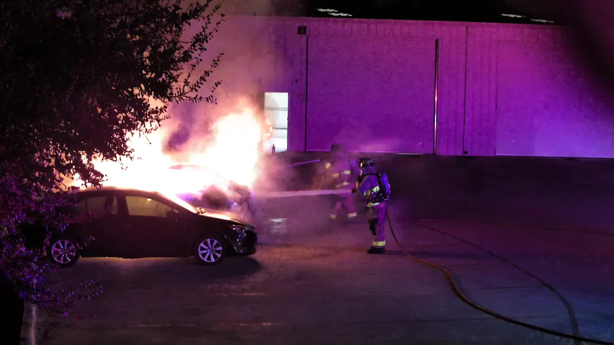 Tower camera at night: firefighters spray a burning car in a parking lot.